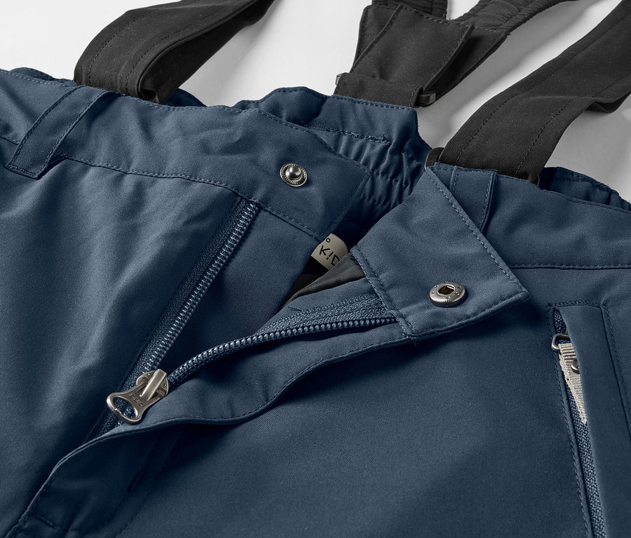 Detailaufnahme einer blauen Hose mit schwarzen Hosenträgern, offenem Reißverschluss und silbernen Knöpfen.