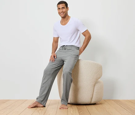 Mann sitzt auf einem beigen Hocker, trägt ein weißes T-Shirt und graue Hosen.