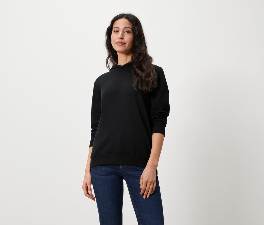 Eine Frau mit langen braunen Haaren steht und trägt einen schwarzen Hoodie und Jeans.