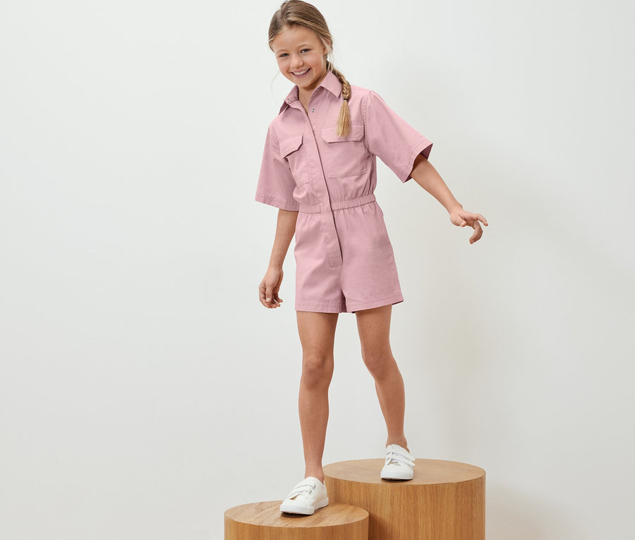 Mädchen steht auf Holzsockeln und trägt einen Kinder-Jumpsuit.