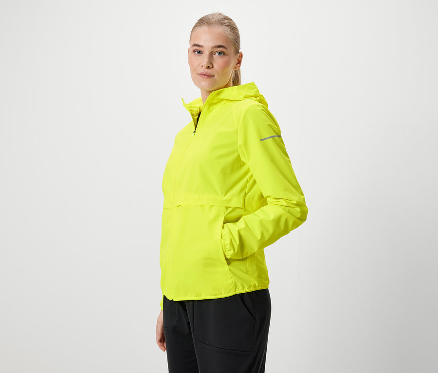 Blonde Frau in neongelber Kapuzenjacke und schwarzen Hosen posiert seitlich.