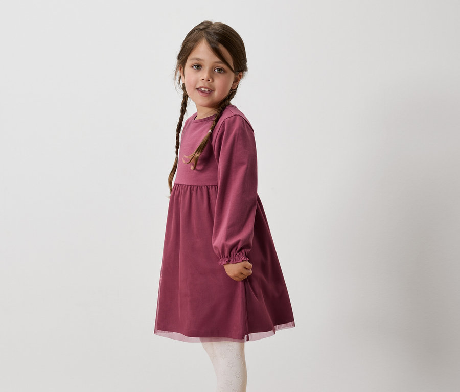 Mädchen mit braunen, zu Zöpfen geflochtenen Haaren trägt ein weinrotes, langärmeliges Kleid und weiße Leggings.