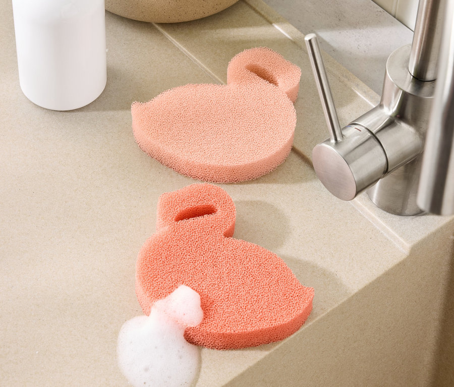 Zwei flamingoförmige Silikonschwämme liegen auf der Arbeitsfläche neben einem Spülbecken, einer davon mit Schaum bedeckt.