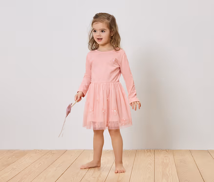 Mädchen mit blonden Haaren trägt ein rosa Kleid und hält einen lila Gegenstand in der Hand.