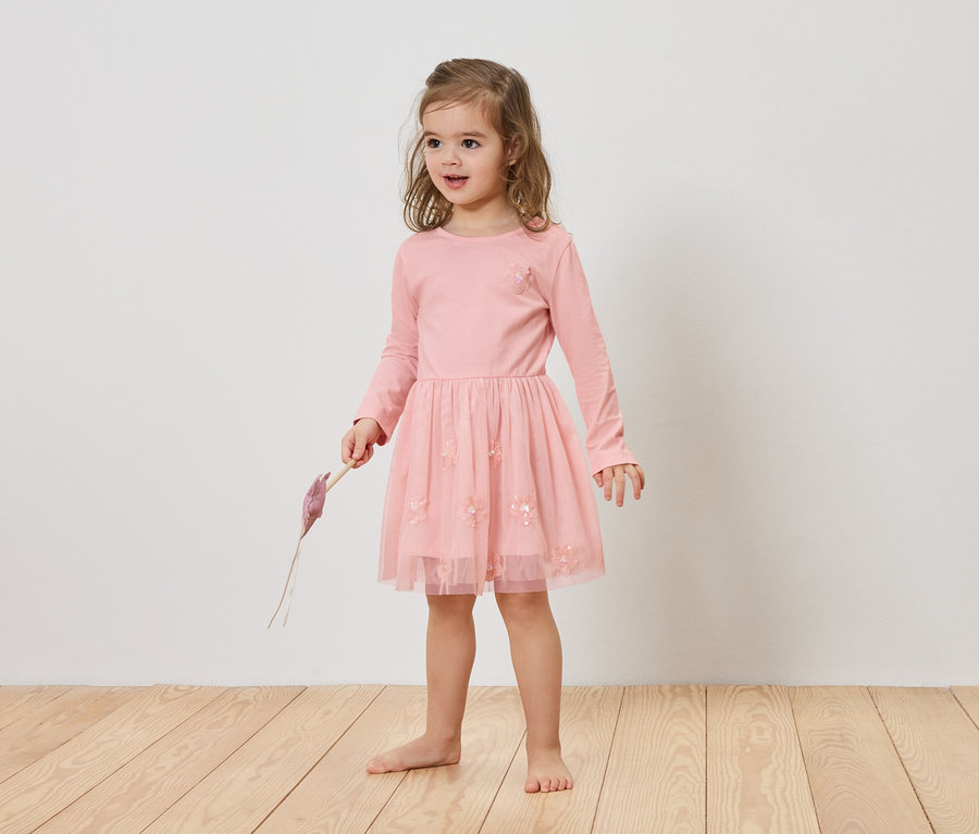 Mädchen mit blonden Haaren trägt ein rosa Kleid und hält einen lila Gegenstand in der Hand.