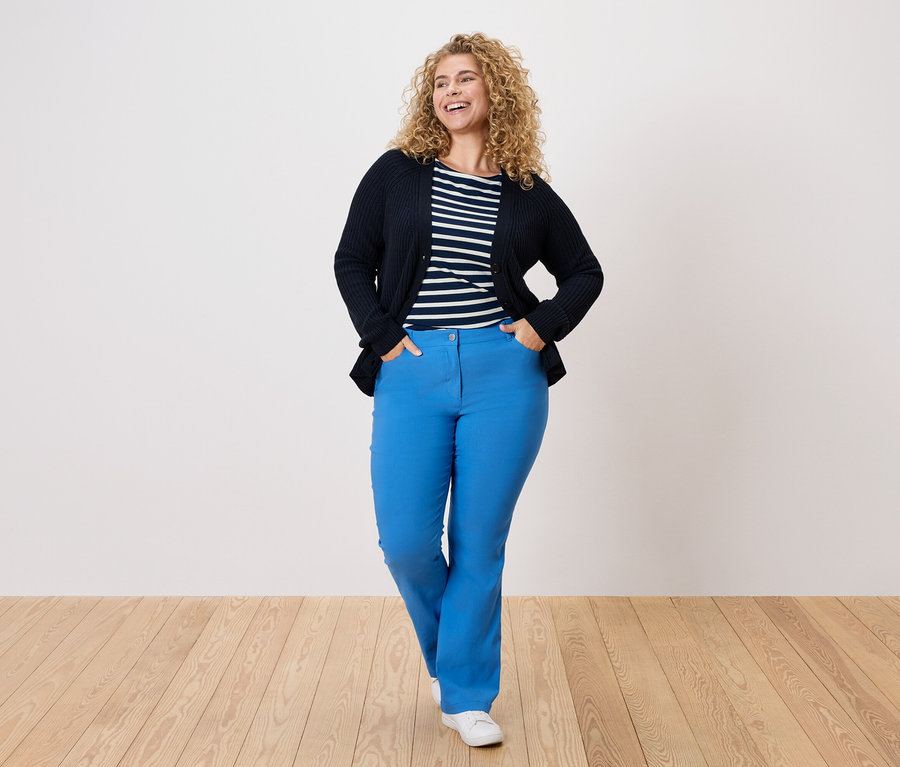 Frau mit lockigem Haar, trägt eine marineblaue Strickjacke, ein gestreiftes Hemd und blaue Hosen.