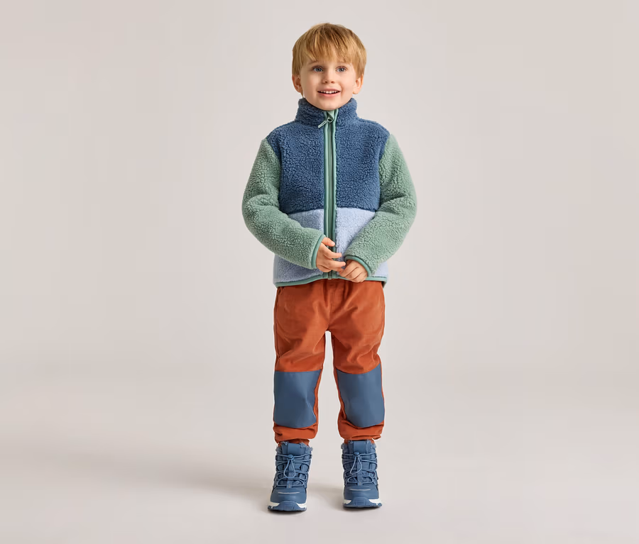 Ein Junge mit blonden Haaren steht vor einem hellen Hintergrund in einer blau-grün-weißen Fleecejacke, orangefarbenen Hosen und blauen Stiefeln.