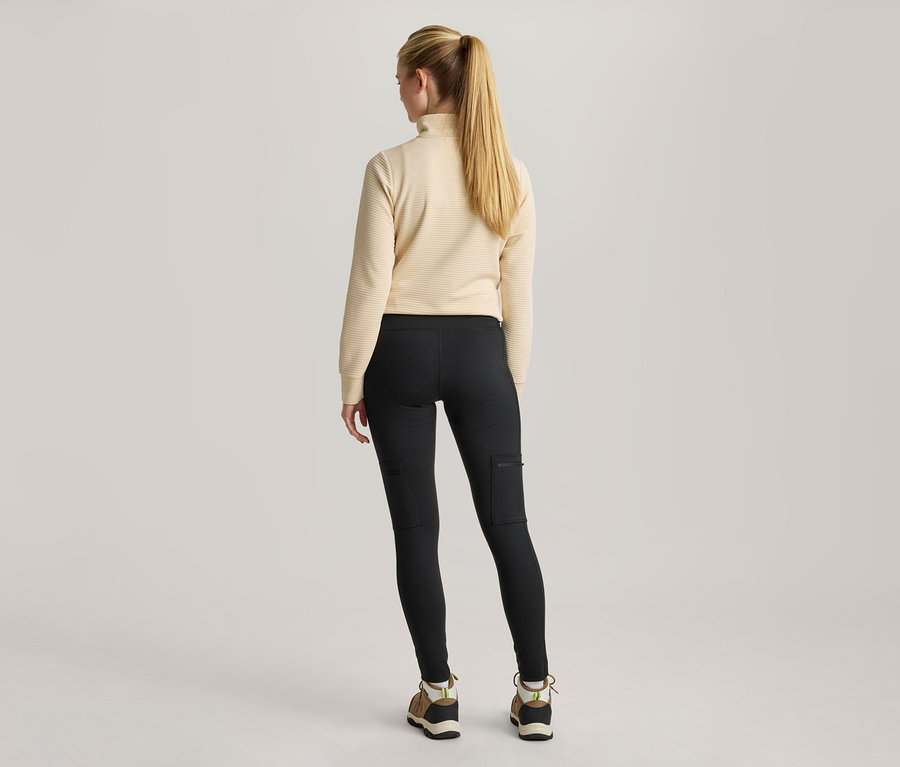 Eine Frau steht mit dem Rücken zum Betrachter und trägt ein cremefarbenes, geripptes Langarmshirt, schwarze Leggings mit Seitentaschen und Wanderschuhe.