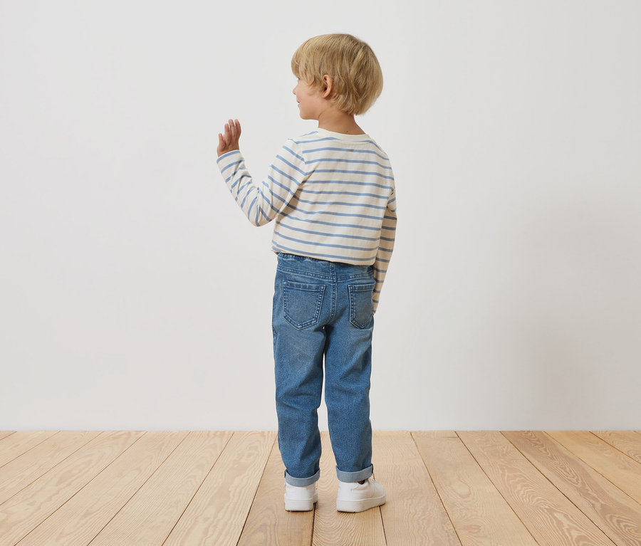 Ein Kind mit blonden Haaren, von hinten gesehen, trägt ein gestreiftes Langarmshirt und Jeans, während es vor einer weißen Wand steht und winkt.