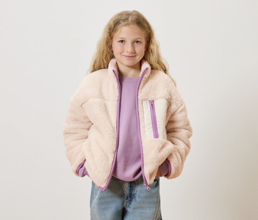 Mädchen mit blonden, lockigen Haaren trägt eine rosa Teddyfelljacke über einem lila Pullover und blauen Jeans.