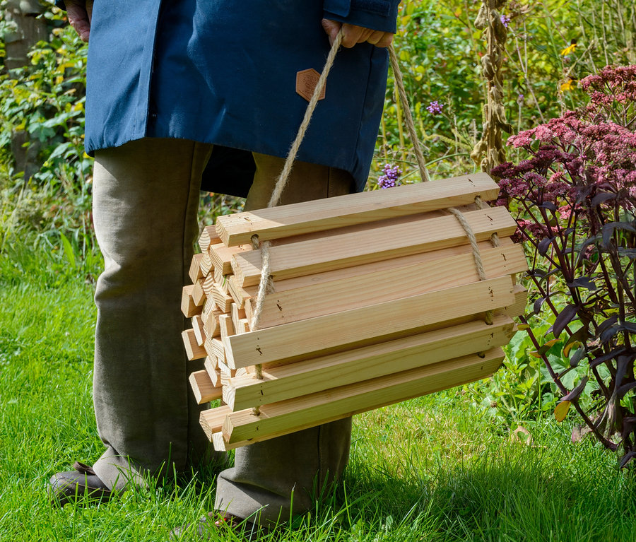 Person trägt eine hölzerne Insektenhotel-Konstruktion mit einem Seil in einem Garten.