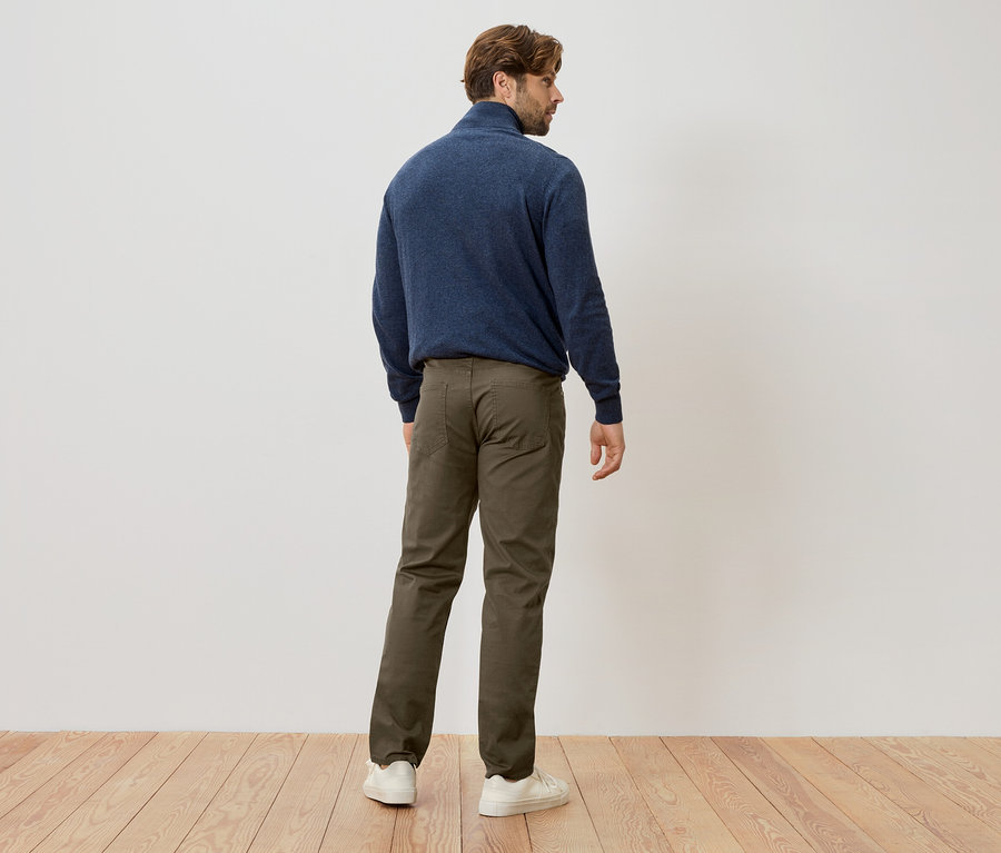 Ein Mann steht mit dem Rücken zur Kamera und trägt einen dunkelblauen Pullover, eine grüne Hose und weiße Turnschuhe.