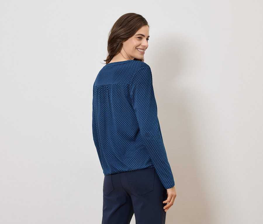 Frau trägt eine blau gemusterte Bluse und dunkle Hose, von hinten gesehen.