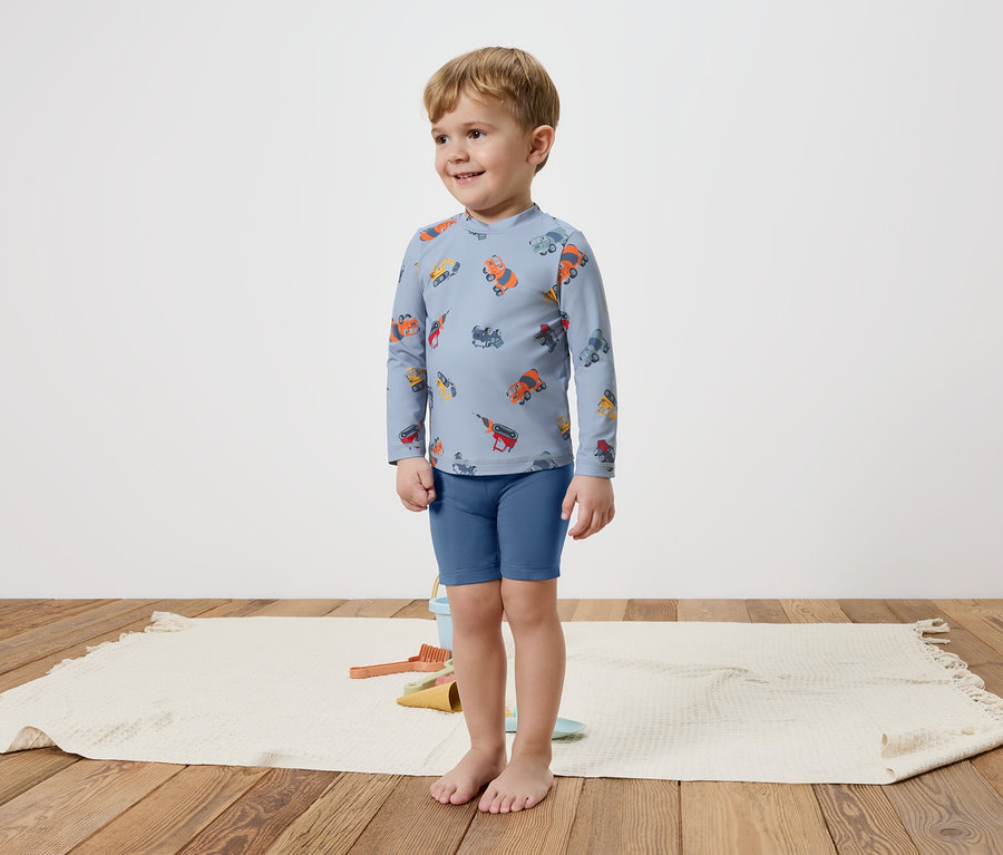 Ein kleiner Junge steht barfuß auf einem Teppich und trägt ein blaues, langärmliges Oberteil mit bunten Fahrzeugen und blaue Shorts.