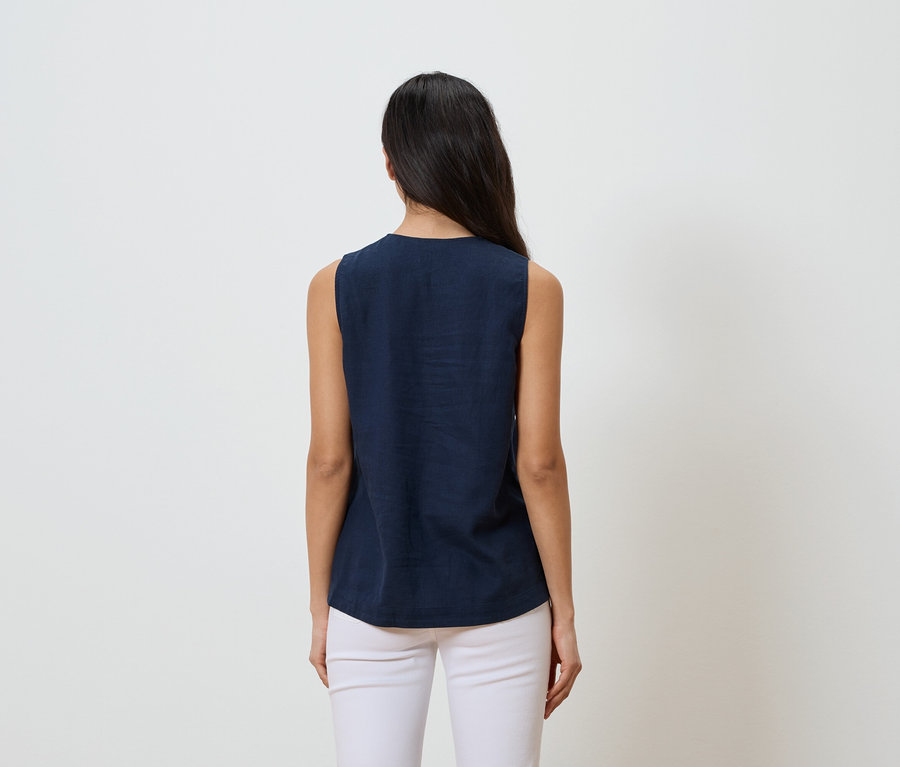 Frau mit dunklen Haaren, von hinten gesehen, trägt ein ärmelloses, dunkelblaues Oberteil und weiße Hosen vor einem weißen Hintergrund.