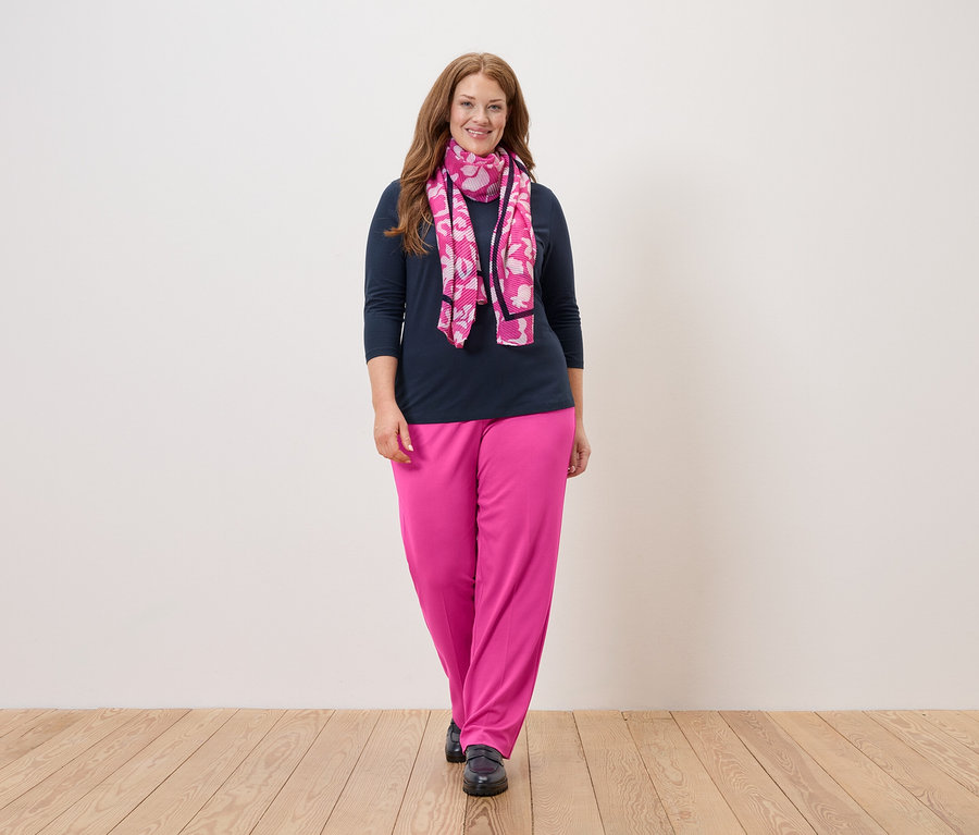 Eine Frau mit langen, roten Haaren steht vor einer weißen Wand und trägt eine marineblaue Bluse, eine pinkfarbene Hose und einen pinkfarbenen Schal mit Blumen.