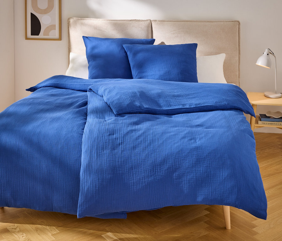 Blaues Bettzeug mit zwei Kissen auf einem Bett mit beigem Kopfteil. Auf dem Nachttisch steht eine Lampe.