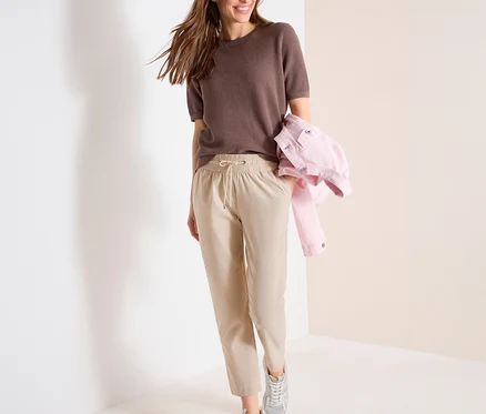 Lächelnde Frau geht, trägt ein braunes Oberteil, eine hellbeige CECIL Joggpants mit Kordelzug und silberne Sneaker, eine rosa Jacke über dem Arm.