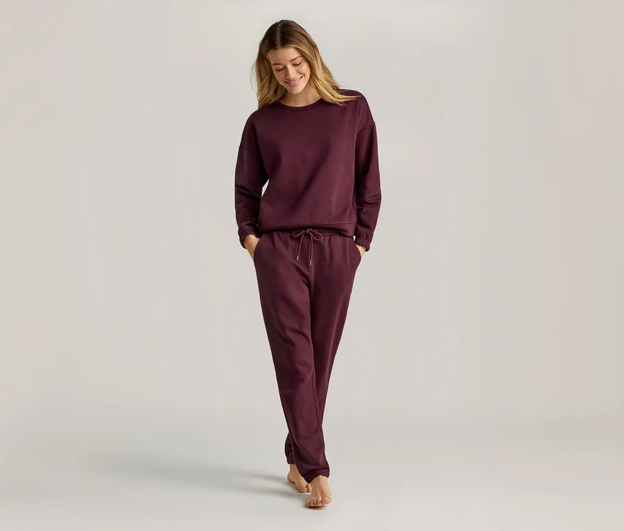 Frau mit langen blonden Haaren, die ein weinrotes, langärmeliges Sweatshirt und eine Jogginghose trägt und barfuß geht.
