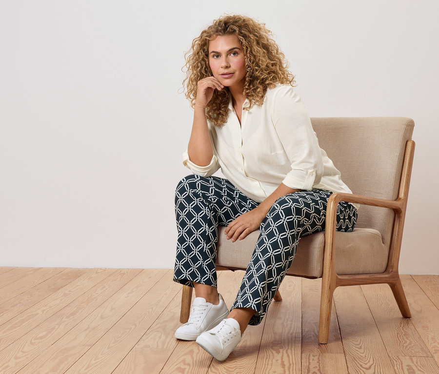 Frau mit lockigen Haaren sitzt in einem Sessel mit gemusterten Hosen und weißen Turnschuhen.