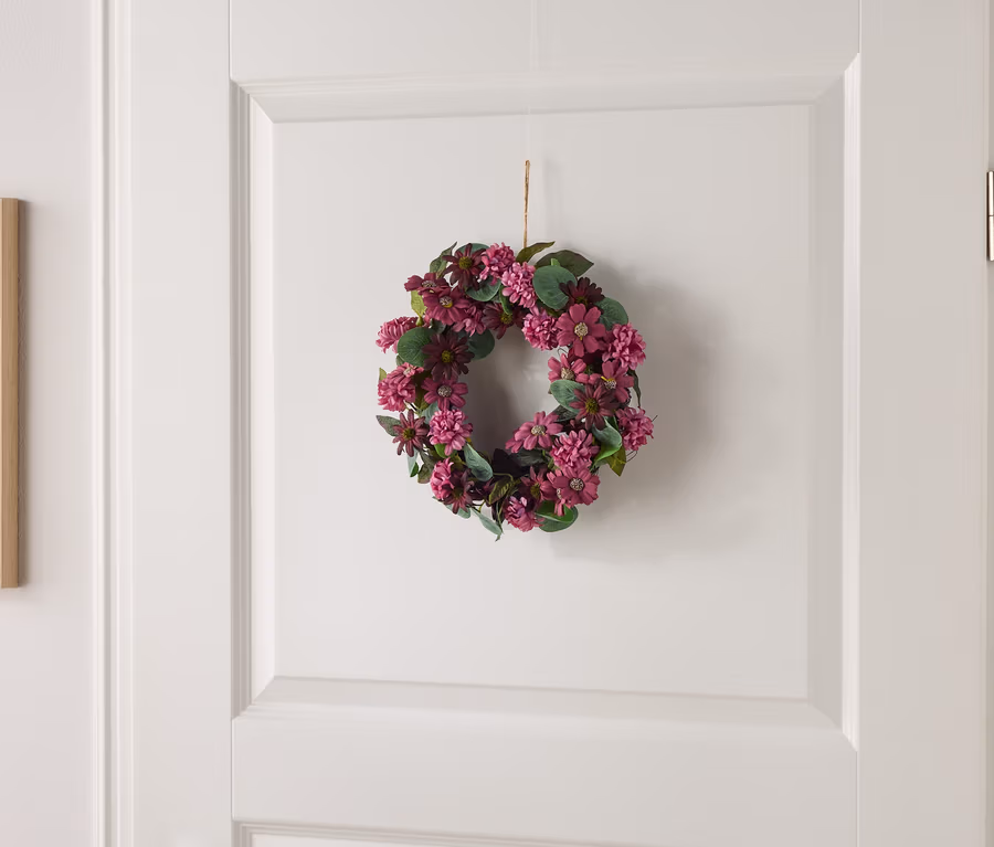 Kleiner Blumenkranz mit lila Blüten und grünen Blättern hängt an einer weißen Tür.