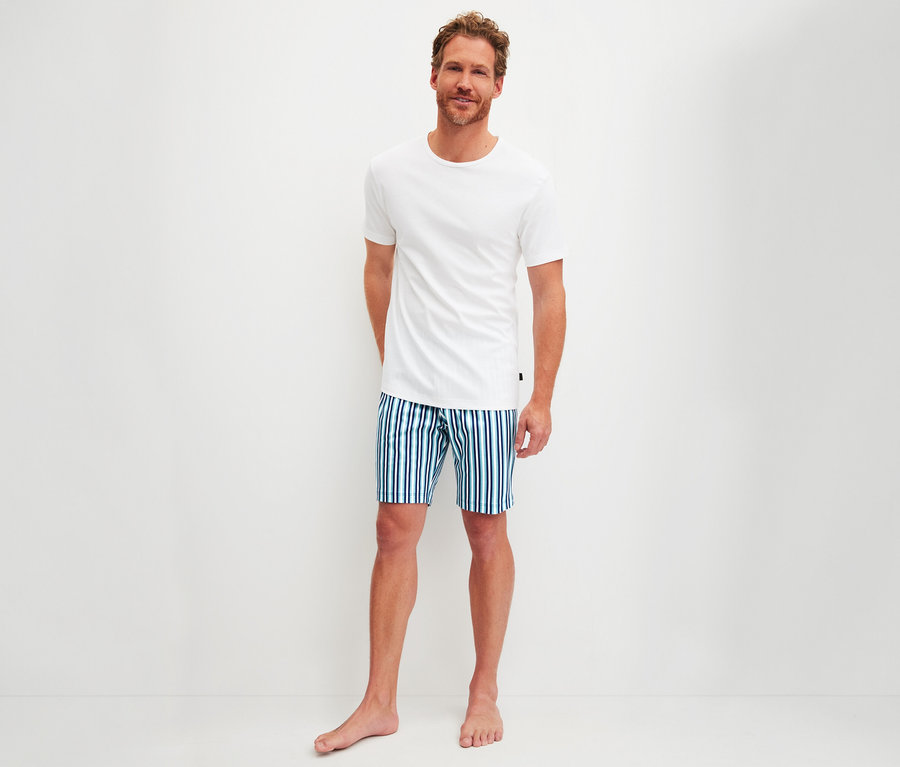 Lächelnder Mann trägt ein weißes T-Shirt und blau-grün gestreifte Shorts aus einem CALIDA Herren-Shorty-Pyjama-Set, barfuß vor weißem Hintergrund stehend.