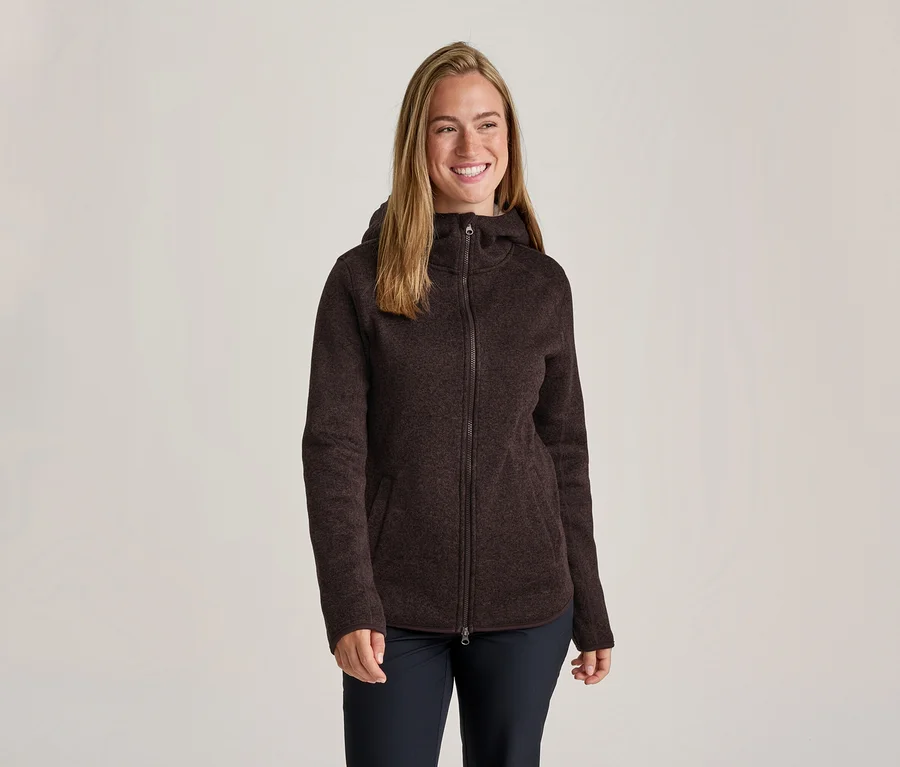 Lächelnde Frau mit langen Haaren trägt eine dunkelbraune, strukturierte Fleecejacke mit Kapuze und Reißverschluss sowie dunkle Hosen.