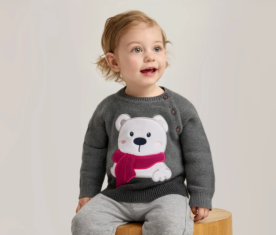 Ein Baby mit blonden Locken sitzt auf einem Holzhocker und trägt einen dunkelgrauen Strickpullover mit einem weißen Eisbären-Motiv, der einen pinken Schal und pinke Wangen hat. Das Baby blickt mit offenem Mund nach oben.