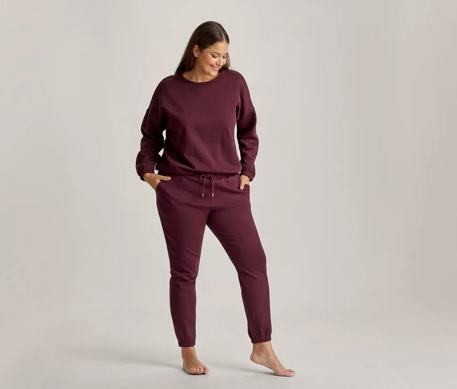 Eine lächelnde Frau steht und blickt nach unten, trägt ein bordeauxrotes Loungewear-Set aus Sweatshirt und Jogginghose. Ihre Hände stecken in den Hosentaschen, und sie ist barfuß.