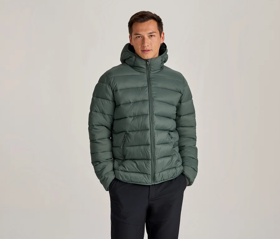 Mann in einer grünen Kapuzen-Steppjacke und dunklen Hosen, der mit den Händen in den Taschen steht.