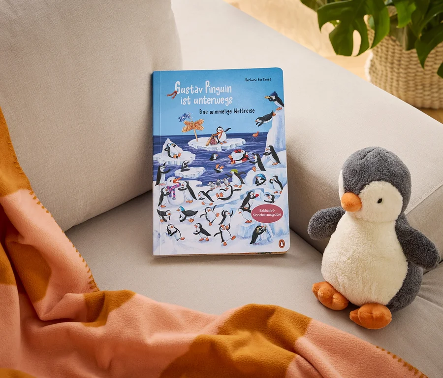 Auf einem Sofa liegen das Kinderbuch "Gustav Pinguin ist unterwegs Eine wimmelige Weltreise" (Exklusive Sonderausgabe), ein Plüschpinguin und eine orange-pfirsichfarbene Streifendecke.