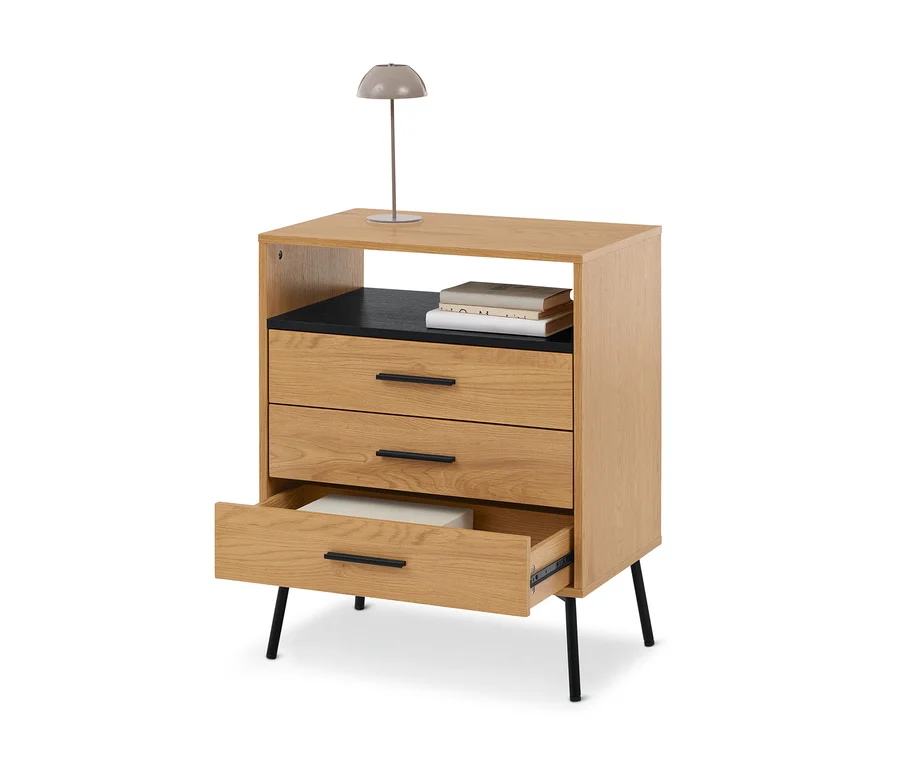 Helle Holzkommode mit drei Schubladen, einer offenen Ablage mit Büchern und einer grauen Lampe oben. Die unterste Schublade ist geöffnet und zeigt einen weißen Gegenstand.
