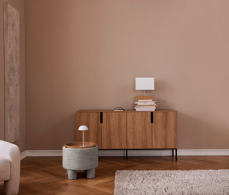 Ein Wohnzimmer mit taupefarbenen Wänden und Fischgrätparkett. Links steht ein beiger Sessel, daneben ein hohes Wandbild. Rechts ein Sideboard aus Holz mit Büchern und einer weißen Lampe darauf. Davor ein runder, gepolsterter Hocker mit Holzplatte und kleiner Lampe. Ein heller Teppich liegt auf dem Boden.