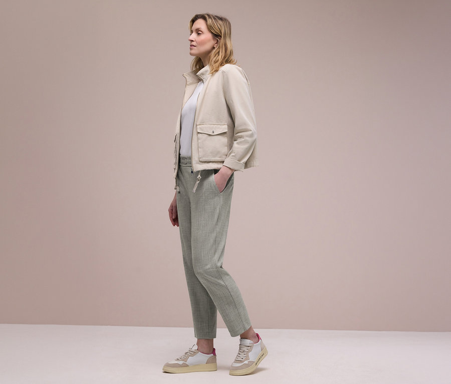Frau im Profil trägt eine beigefarbene Jacke, ein weißes Oberteil und hellgrüne Street One Joggpants, eine Hand in der Tasche. Sie trägt beige-weiße Sneaker.
