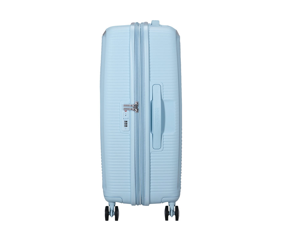 Ein mittelgroßer American Tourister Soundbox Spinner Hartschalenkoffer in Pastellblau, von der Seite stehend. Er hat eine gerippte Textur, einen seitlichen Tragegriff, ein Zahlenschloss und vier Rollen.