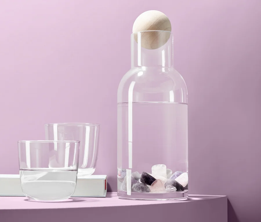 Eine Glaskaraffe mit Wasser und Wassersteine, dazu zwei Gläser, vor einem lila Hintergrund.