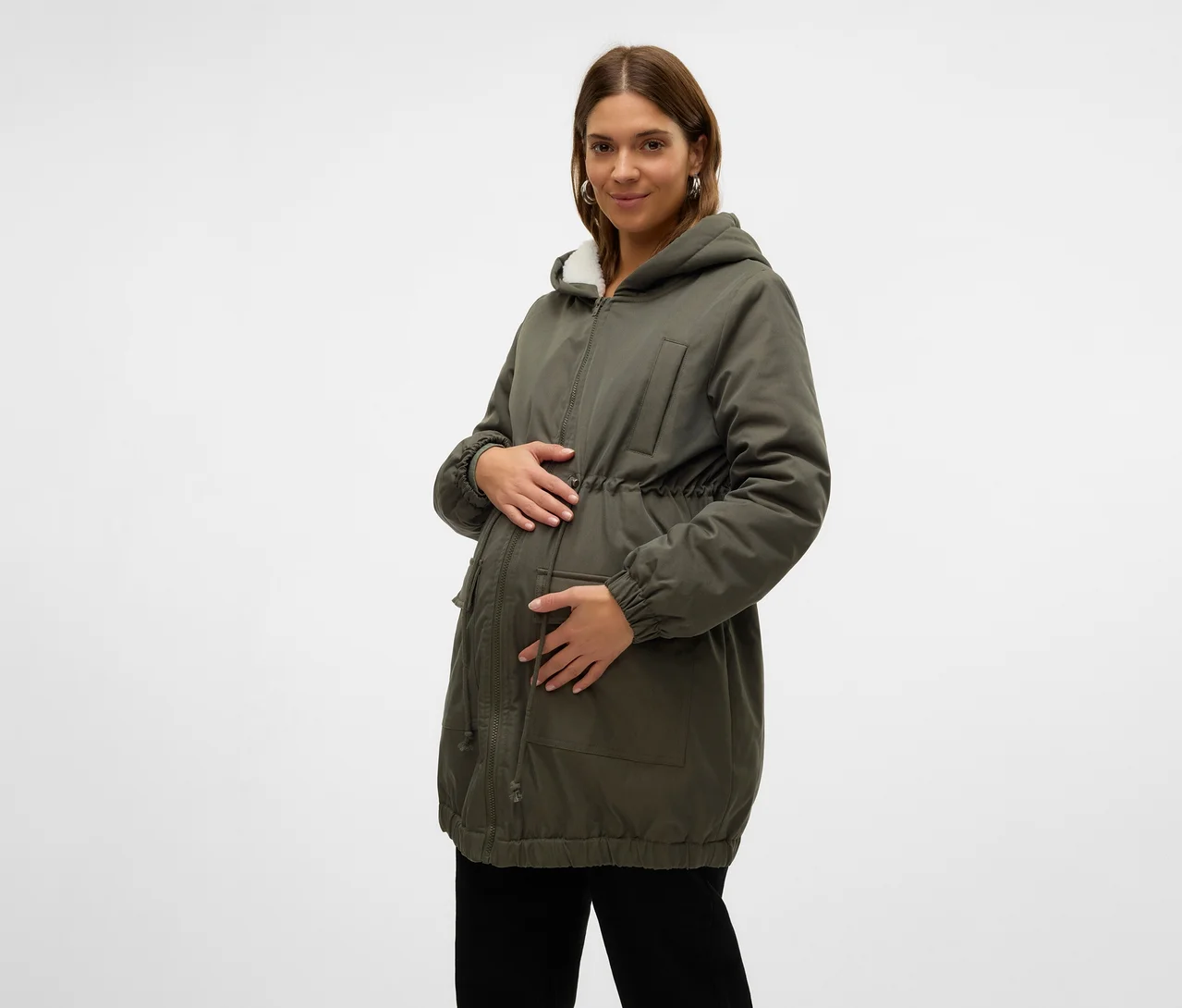 Schwangere Frau in einem grünen Parka mit weißem Kunstfell gefütterter Kapuze und Kordelzug in der Taille, die ihren Bauch hält. Sie trägt auch eine schwarze Hose.