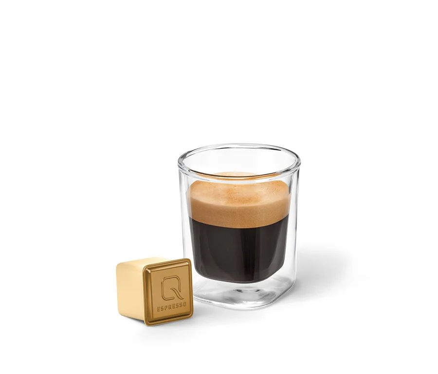 Ein Glas Qbo Flavoured Espresso Zimtstern mit Crema steht neben einer goldenen, würfelförmigen Qbo Espresso Kapsel.