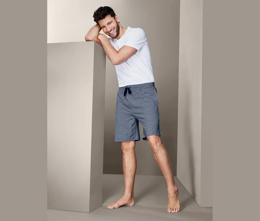 Mann lehnt an einer Wand, trägt ein weißes T-Shirt und Schlafshorts.