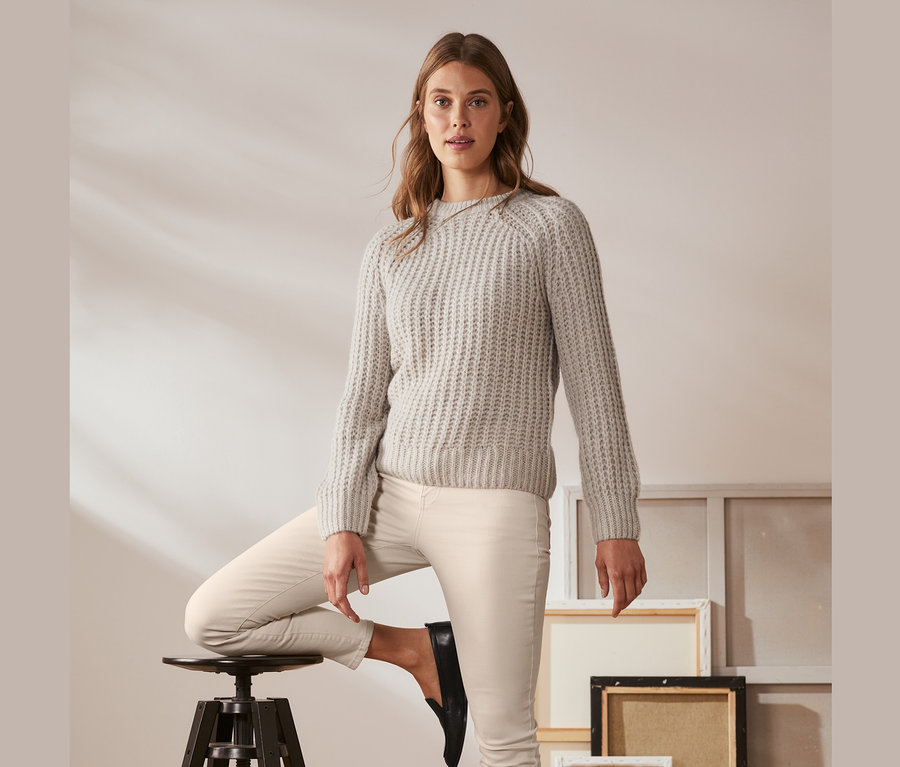 Eine Frau posiert mit einem Bein auf einem Barhocker, sie trägt einen cremefarben melierten Grobstrick-Pullover mit Wolle.