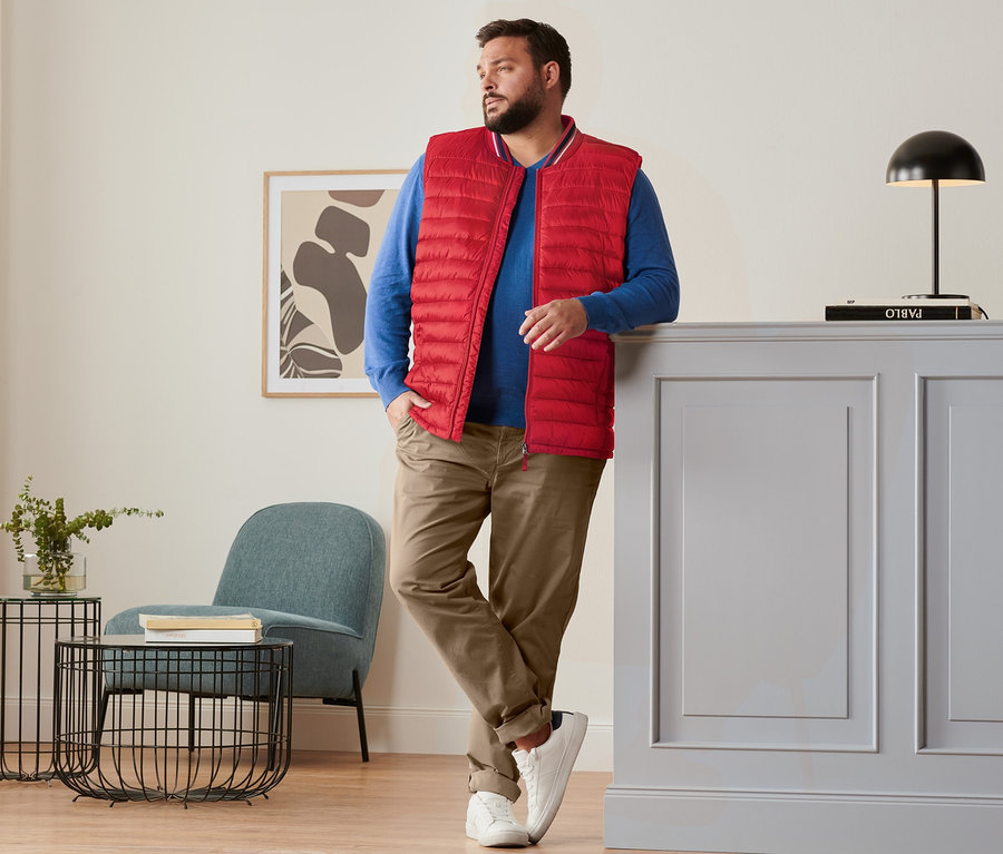 Mann lehnt an Kommode, trägt rote Steppweste, royalblauen Leinen-Pullover, camelfarbene Chino und Sneaker.