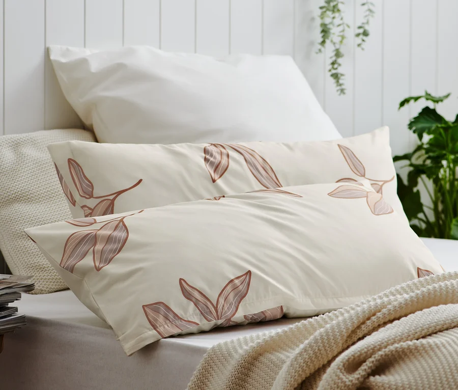 Zwei beige Renforcé-Kopfkissenbezüge mit Blattmuster liegen auf einem Bett.