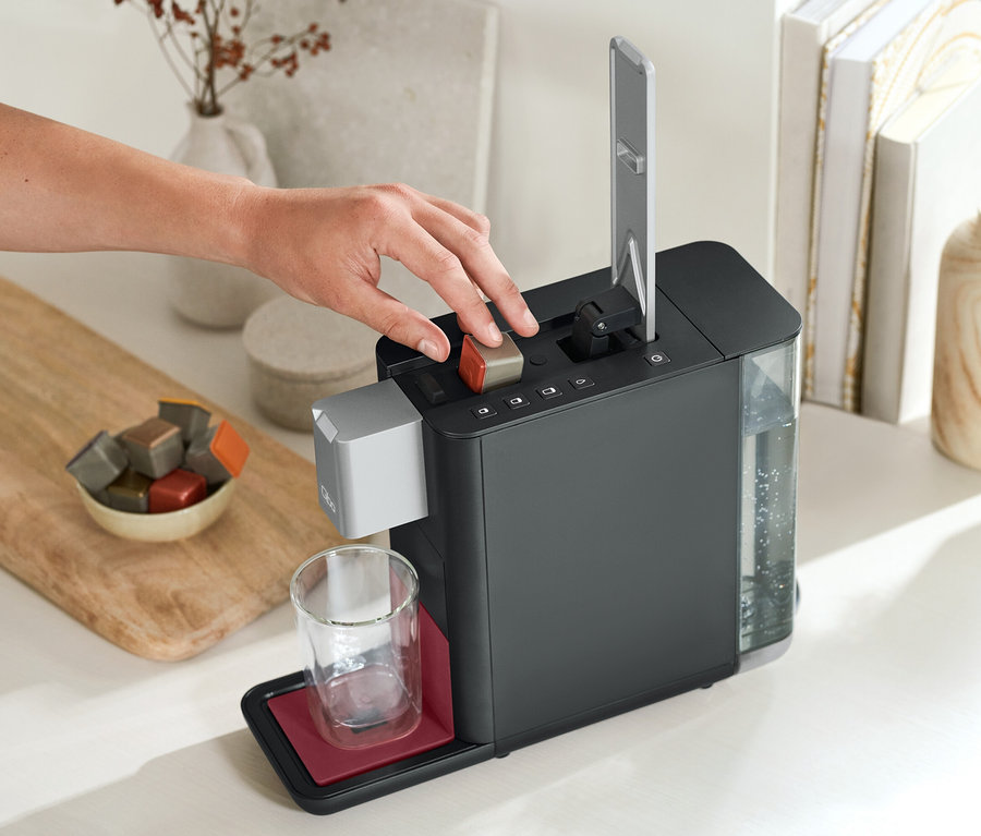 Eine Hand drückt einen Knopf auf einer Qbo ESSENTIAL Dark Berry Kaffeemaschine. Daneben stehen ein Glas und eine Schale mit Kapseln.