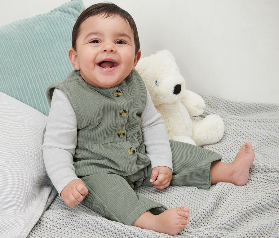 Ein lächelndes Baby sitzt auf einer grauen Strickdecke neben einem Teddybär und trägt eine Baby-Cordlatzhose.