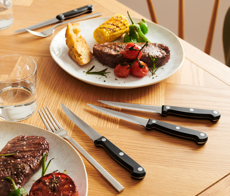 Auf einem Holztisch stehen zwei Teller mit Rindersteak, Mais und Tomaten. Neben den Tellern liegen silbernes Besteck und ein Set Steakmesser.