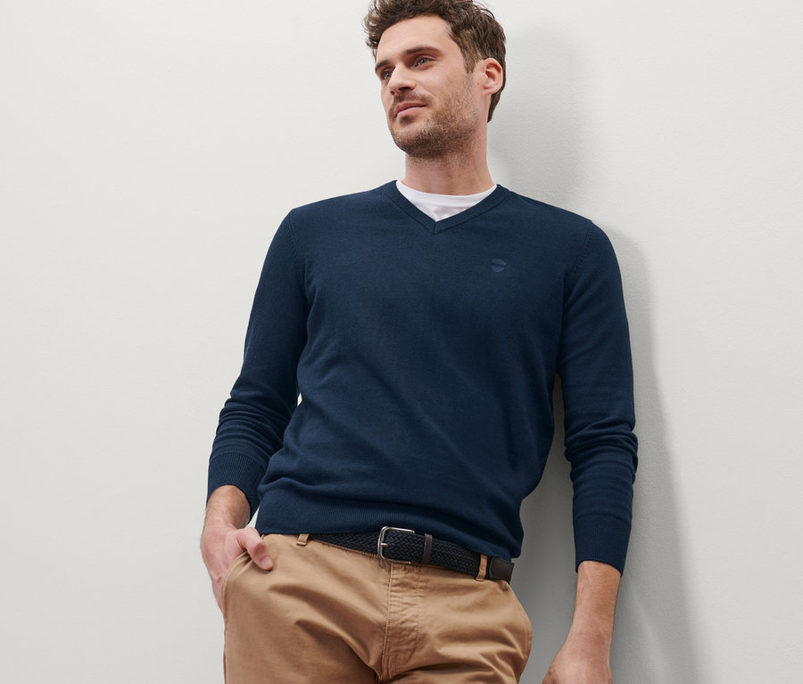 Mann lehnt an Wand und trägt marineblauen Pullover mit V-Ausschnitt.