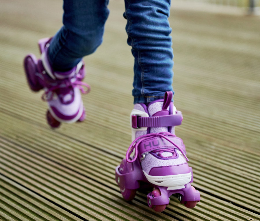 Kind in Jeans und Jacke läuft auf lila HUDORA Rollschuhen »My First Quad« auf einem Holzdeck.