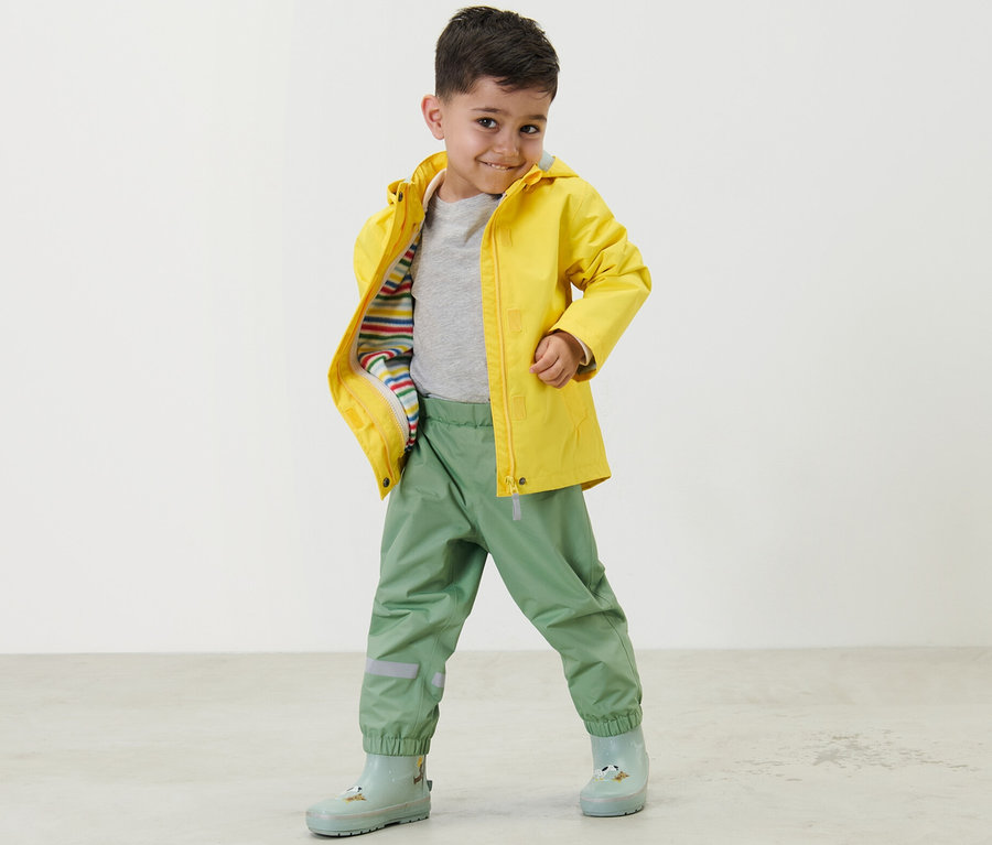 Ein Junge posiert in grasgrünen Kinder-Regenhosen und einer gelben Regenjacke.
