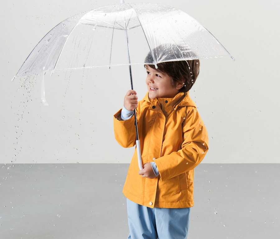 Ein Junge hält einen durchsichtigen Regenschirm und trägt eine korngelbe Kleinkind-Thermo-Regenjacke.