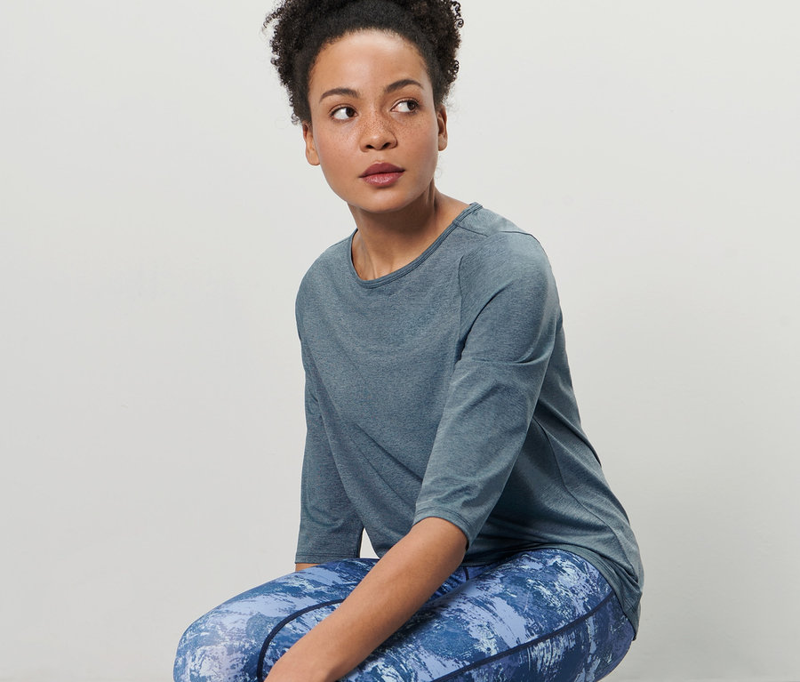 Frau in dunkelblauem Funktionsshirt mit 3/4-Ärmeln und blauen gemusterten Leggings dehnt sich.